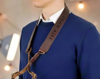 May include: A brown leather camera strap with the text "JOHN ALEX" in white lettering. The strap is worn over a navy blue sweater and white collared shirt. A black camera is held in front of the person.