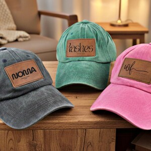 Può includere: Tre cappellini da baseball grigi, verdi e rosa, ciascuno con una toppa rettangolare. Il cappellino grigio recita "NONNA", quello verde "lashes" e quello rosa "wcf". Tutti i cappellini sono su una superficie di legno.
