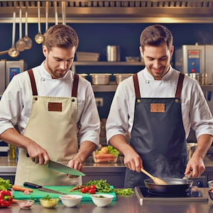 May include: Two chefs in a commercial kitchen, wearing white coats and aprons with brown leather straps. One chef is chopping vegetables with a large knife, while the other is cooking in a pan. The kitchen has stainless steel appliances and various cooking utensils.
