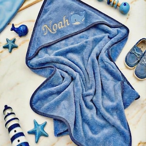 May include: A blue hooded baby towel with the name "Noah" embroidered in gold, along with a whale illustration. The towel is surrounded by nautical-themed decorations, including a toy rattle, shoes, a lighthouse, and starfish.