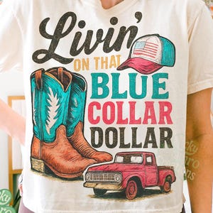 Puede incluir: Camiseta color crema con un diseño gráfico que presenta botas de vaquero, una gorra de béisbol y una camioneta roja. El texto dice "Livin' on that Blue Collar Dollar". La camiseta es una descarga digital.