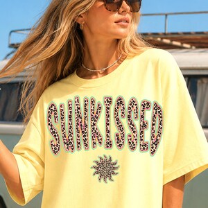 以下が含まれることがあります： ヒョウ柄のフォントで「SUNKISSED」の文字と太陽のグラフィックが入った黄色のTシャツ。ビーチで、背景にヴィンテージのバンがある画像です。