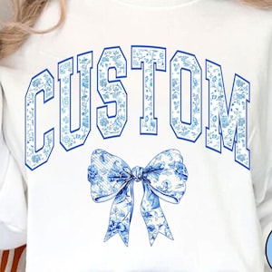 May include: White sweatshirt with a blue and white floral pattern spelling "CUSTOM" in a collegiate font. A blue and white floral bow is centred below the text.