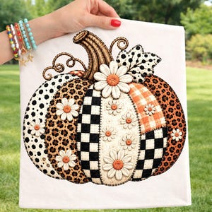 Puede incluir: Un cuadrado de tela blanca con una ilustración de una calabaza grande compuesta por varios parches estampados, incluyendo cuadros blancos y negros, estampado de leopardo y cuadros naranjas, decorada con margaritas blancas.