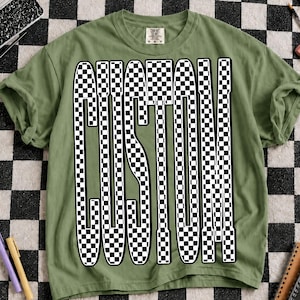 May include: An olive green short-sleeve t-shirt featuring the word CUSTOM printed in a large, bold, black and white checkered pattern. The letters are elongated and vertically oriented, centered on the front of the garment with a crew neckline.