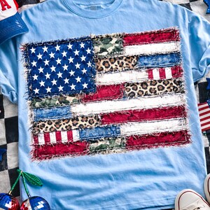 Op de afbeelding: Lichtblauw T-shirt met een patchwork Amerikaans vlagontwerp. De vlag bevat denim, luipaardprint en rood-witte strepen. Een rood-wit-blauwe baseballcap met "USA" is zichtbaar. Patriotisch thema.