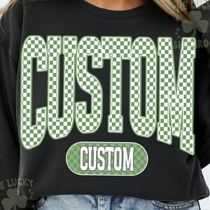 May include: Black sweatshirt with the word "CUSTOM" in large, green and white checkered letters across the front. A smaller, oval-shaped "CUSTOM" design is below the main text. The sweatshirt is a casual style.