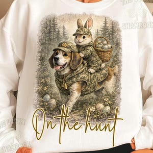 May include: White sweatshirt featuring a beagle dog wearing a camouflage outfit, carrying a bunny wearing a matching outfit and a basket of eggs. The text "On the hunt" is written in gold script.