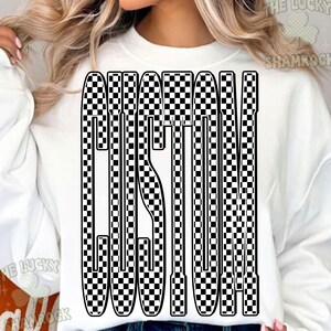 May include: White sweatshirt featuring the word "CUSTOM" in a black and white chequered pattern. The design is in a vertical, block letter style. The sweatshirt has a crew neck and long sleeves.