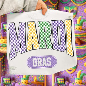 May include: White sweatshirt with the words "MARDI GRAS" in purple, green, and yellow chequered letters. The word "GRAS" is in a purple oval. The sweatshirt is held up by a hand.