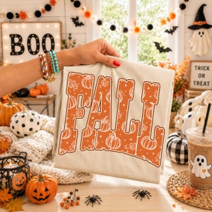 May include: A beige t-shirt featuring the word FALL in large, bold, orange letters. The letters are filled with a white line-art pattern of pumpkins, leaves, and stars. The shirt is held up against a background of various Halloween decorations.