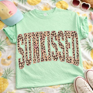 May include: A light green t-shirt featuring the word SUNKISSED in a large, bold, arched font with a brown and black leopard print pattern. The letters are outlined in a thin pink border, creating a distinct graphic design on the front of the shirt.