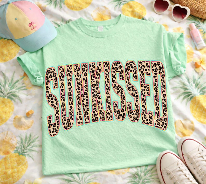 May include: A light green t-shirt featuring the word SUNKISSED in a large, bold, arched font with a brown and black leopard print pattern. The letters are outlined in a thin pink border, creating a distinct graphic design on the front of the shirt.