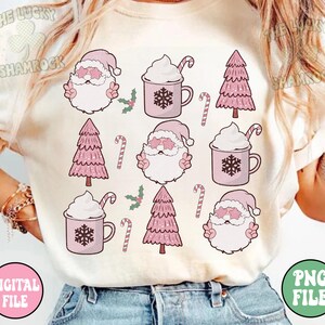 K&ouml;nnte beinhalten: Ein wei&szlig;es T-Shirt mit einem rosa-wei&szlig;en Weihnachtsmotiv. Das Design zeigt den Weihnachtsmann, Weihnachtsb&auml;ume und Tassen mit hei&szlig;er Schokolade mit Schlagsahne.