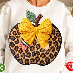 May include: White sweatshirt featuring a large, textured apple design. The apple has a leopard print pattern, a yellow bow, a green leaf, and a brown stem. A small pencil and a pink bow are also included in the design.