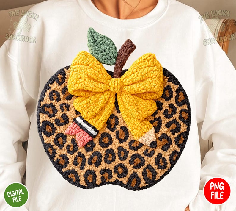 May include: White sweatshirt featuring a large, textured apple design. The apple has a leopard print pattern, a yellow bow, a green leaf, and a brown stem. A small pencil and a pink bow are also included in the design.