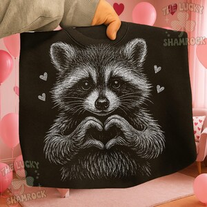 Peut inclure: Sweat-shirt noir avec une illustration blanche d'un raton laveur formant un c&oelig;ur avec ses mains. Le raton laveur a l'air duveteux, avec de petits c&oelig;urs dispers&eacute;s. Le texte "THE LUCKY SHAMROCK" est visible en arri&egrave;re-plan.