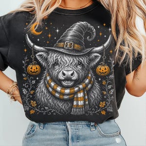 May include: A black crewneck sweatshirt featuring a graphic illustration of a Highland cow wearing a witch hat and a striped scarf. The cow is flanked by two hanging jack-o'-lanterns, with a crescent moon, stars, and autumn leaves surrounding it.