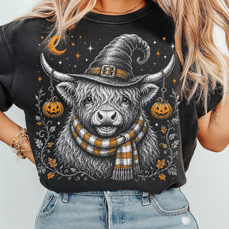 May include: A black crewneck sweatshirt featuring a graphic illustration of a Highland cow wearing a witch hat and a striped scarf. The cow is flanked by two hanging jack-o'-lanterns, with a crescent moon, stars, and autumn leaves surrounding it.
