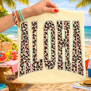 May include: Cream-colored t-shirt with the word "ALOHA" in large, bold letters. The letters feature a leopard print design with pink and teal accents. The shirt is held up against a beach backdrop.