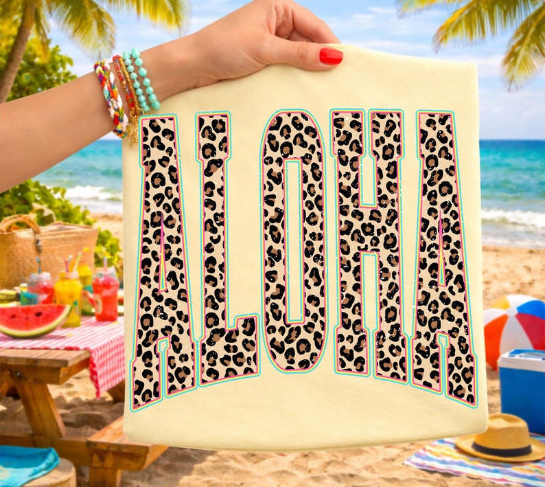May include: Cream-colored t-shirt with the word "ALOHA" in large, bold letters. The letters feature a leopard print design with pink and teal accents. The shirt is held up against a beach backdrop.