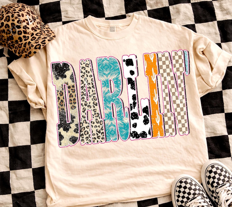 May include: Cream-colored t-shirt with the word "DARLIN" in a colorful, patterned design. The letters feature animal prints, turquoise, and checkered patterns. A leopard print hat and checkered sneakers are also visible.