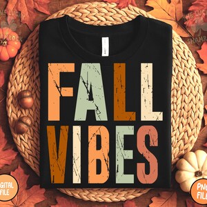 May include: Black t-shirt with the words "FALL VIBES" in a distressed, multi-colored font. The shirt is surrounded by fall-themed decorations, including pumpkins, leaves, and acorns. The shirt is displayed on a woven mat.