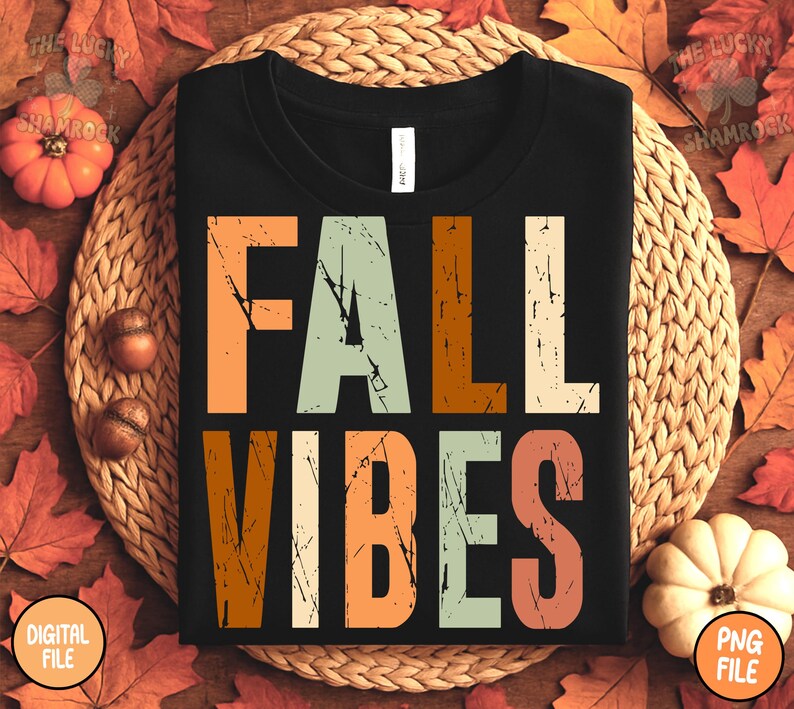 May include: Black t-shirt with the words "FALL VIBES" in a distressed, multi-colored font. The shirt is surrounded by fall-themed decorations, including pumpkins, leaves, and acorns. The shirt is displayed on a woven mat.