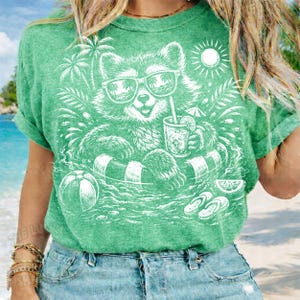 May include: Green t-shirt featuring a white graphic of a raccoon wearing sunglasses and floating in a pool with a drink. The design includes a beach ball, palm trees, and a sun.