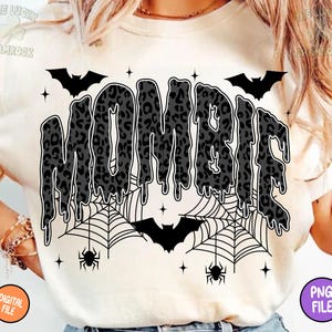 May include: Cream-colored t-shirt with the word "MOMBIE" in a dripping, leopard-print design. The design includes bats, spiderwebs, spiders, and stars, creating a Halloween theme. The shirt also has the text "DIGITAL FILE" and "PNG FILE".
