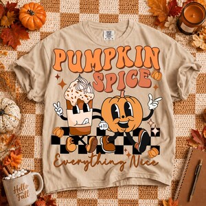 May include: A beige t-shirt featuring a graphic print with the text PUMPKIN SPICE and Everything Nice. The design includes a cartoon coffee cup and a pumpkin with arms and legs, set against a black and white checkered pattern with stars.