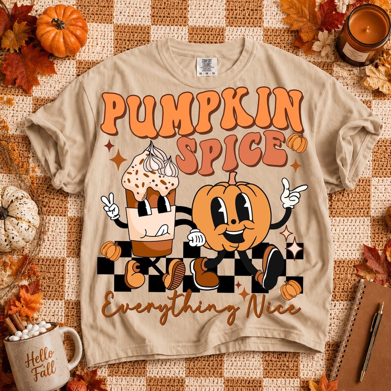 May include: A beige t-shirt featuring a graphic print with the text PUMPKIN SPICE and Everything Nice. The design includes a cartoon coffee cup and a pumpkin with arms and legs, set against a black and white checkered pattern with stars.