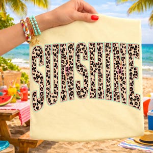 May include: A light yellow t-shirt with the word "SUNSHINE" in large, leopard print letters. The letters have a light blue outline. The shirt is held up by a person with a red manicure, with a beach scene in the background.