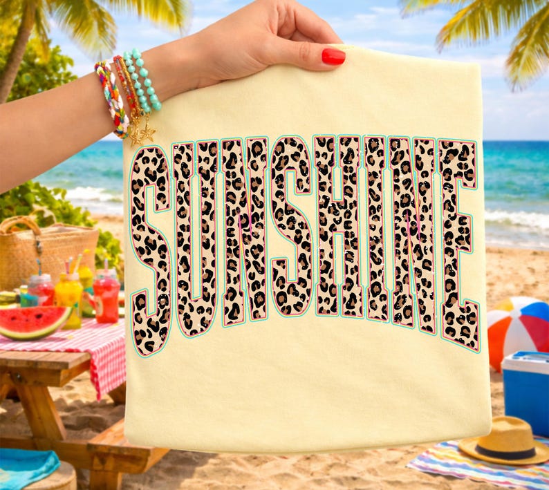 May include: A light yellow t-shirt with the word "SUNSHINE" in large, leopard print letters. The letters have a light blue outline. The shirt is held up by a person with a red manicure, with a beach scene in the background.