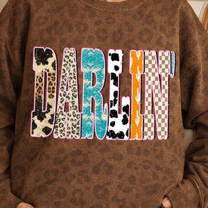 May include: Brown sweatshirt with the word "DARLIN" in a colorful, patterned design. The letters feature animal print, turquoise, black and white cow spots, orange cactus, and a checkered pattern. The sweatshirt has a leopard print pattern.