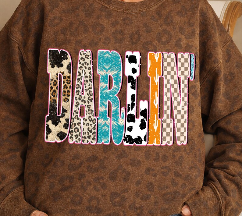 May include: Brown sweatshirt with the word "DARLIN" in a colorful, patterned design. The letters feature animal print, turquoise, black and white cow spots, orange cactus, and a checkered pattern. The sweatshirt has a leopard print pattern.