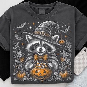 May include: A charcoal gray t-shirt featuring a graphic illustration of a raccoon wearing a witch hat and a polka-dot bow tie. The raccoon holds a jack-o'-lantern filled with candy corn, surrounded by bats, orange stars, and decorative foliage.