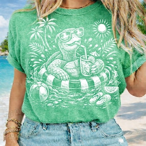 May include: Green t-shirt featuring a white graphic of a turtle wearing sunglasses and floating in a pool with a drink. The design includes a beach ball, palm trees, and a sun, evoking a summer theme.