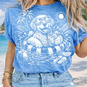 May include: Blue t-shirt with a white graphic of a dog wearing sunglasses and floating in a pool with a drink. The design includes palm trees, a beach ball, and the sun. The shirt has rolled-up sleeves.