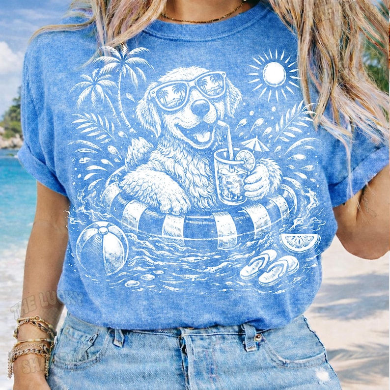 May include: Blue t-shirt with a white graphic of a dog wearing sunglasses and floating in a pool with a drink. The design includes palm trees, a beach ball, and the sun. The shirt has rolled-up sleeves.