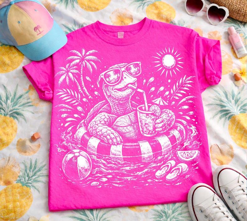 May include: Vibrant pink t-shirt with a white graphic of a turtle wearing sunglasses and floating in a pool with a drink. The design includes palm trees, a beach ball, and a sun. A blue and pink hat is visible.