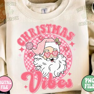 May include: Cream-colored sweatshirt with a retro graphic of Santa Claus wearing heart-shaped sunglasses and a pink Santa hat. The text "Christmas Vibes" is in pink, with a checkered background and star accents. A digital file is available.