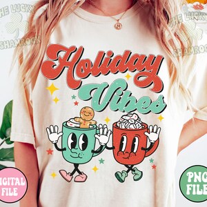 May include: Cream-colored t-shirt with the words "Holiday Vibes" in red and teal retro font. Two cartoon mugs with faces and arms are featured below the text, one with a gingerbread man and the other with marshmallows. Stars and the words "Digital File" and "PNG File" are also visible.