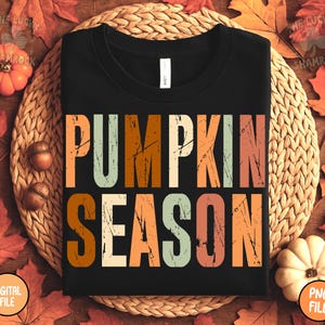 May include: Black t-shirt with the words "PUMPKIN SEASON" in large, distressed, colorful letters. The shirt is surrounded by fall leaves, pumpkins, and acorns, creating a seasonal autumn theme.
