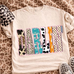 May include: Cream-colored t-shirt with the word "DARLIN" in a colorful, patterned design. The patterns include leopard print, turquoise, cow print, orange cactus, and checkered patterns. A checkered hat and shoes are also visible.
