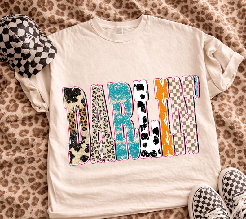 May include: Cream-colored t-shirt with the word "DARLIN" in a colorful, patterned design. The patterns include leopard print, turquoise, cow print, orange cactus, and checkered patterns. A checkered hat and shoes are also visible.
