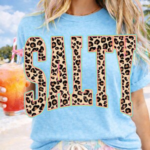 May include: Light blue t-shirt with the word "SALTY" in large, leopard print letters. The letters have a pink outline and a turquoise border. The person is holding a tropical drink with a pink umbrella.