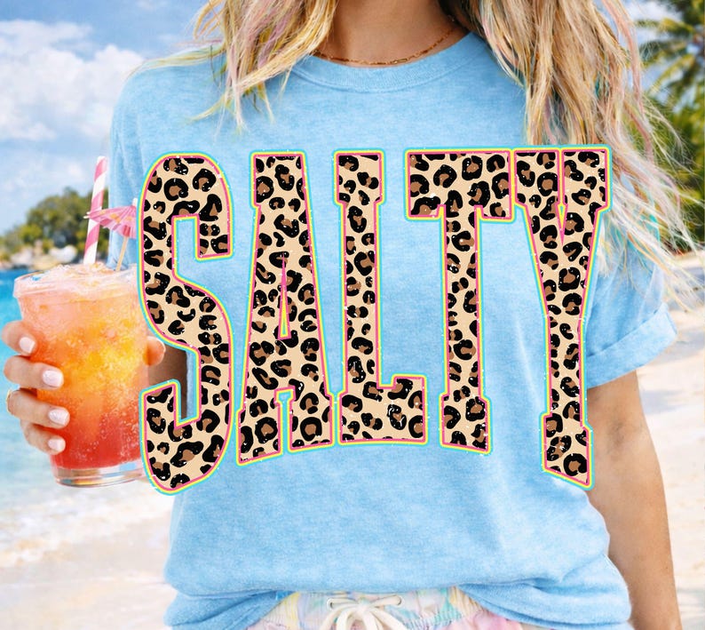 May include: Light blue t-shirt with the word "SALTY" in large, leopard print letters. The letters have a pink outline and a turquoise border. The person is holding a tropical drink with a pink umbrella.
