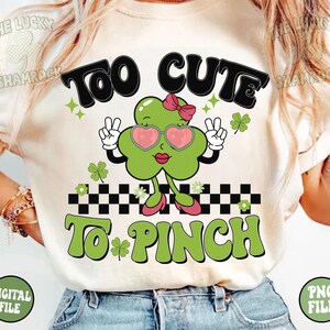 Peut inclure: Un t-shirt blanc avec un tr&egrave;fle vert portant des lunettes de soleil et un n&oelig;ud rose. Le tr&egrave;fle l&egrave;ve deux doigts en signe de paix. Le texte "Too Cute To Pinch" est &eacute;crit en lettres vertes et noires.