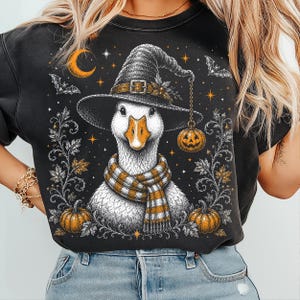 May include: A black crewneck sweatshirt featuring a graphic illustration of a white goose wearing a pointed witch hat and a striped orange and white scarf. The design includes a hanging jack-o'-lantern, bats, a crescent moon, and pumpkins.